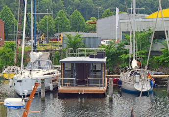 Huslysbåd Leni fortøjet mellem to sejlbåde ved en havn i Tyskland omgivet af grønne træer.