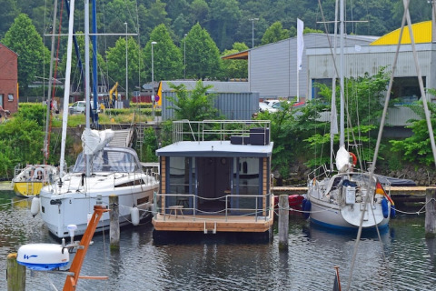 Woonboot Leni aangemeerd tussen twee zeilboten in een Duitse haven met groene bomen op de achtergrond.