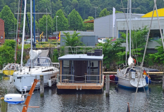 Woonboot Leni aangemeerd tussen twee zeilboten in een Duitse haven met groene bomen op de achtergrond.