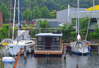 Le bateau-maison Leni amarré entre deux voiliers dans un port allemand entouré d’arbres verts.