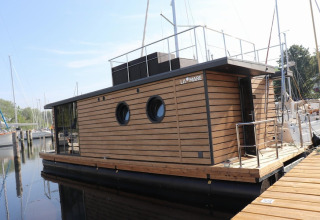 Moderne husbåd kaldet Houseboat «Leni» ved en havn i Tyskland, med træbeklædning og runde vinduer.