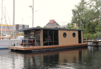 Het moderne woonboot Houseboat Lina ligt aangemeerd in een haven, omgeven door boten en groen.