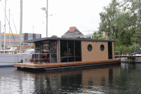 The modern Houseboat Lina is docked in a marina, surrounded by sailboats and waterside greenery.
