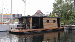 Et moderne husbåd ved navn Houseboat Lina ligger fortøjet i en havn omgivet af andre både og natur.