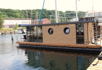 Woonboot «Lina» aangemeerd in een haven in Duitsland, met groene heuvels en boten op de achtergrond.
