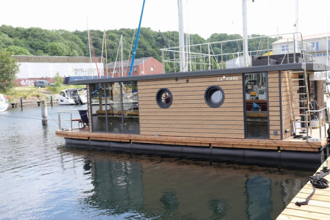 Houseboat «Lina» docked at a marina in Germany, surrounded by lush green hills and nearby boats.