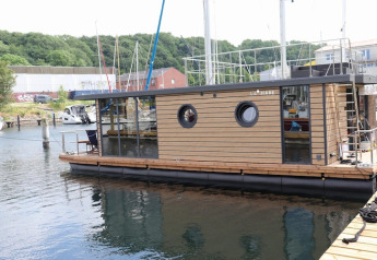 Casa flotante «Lina» amarrada en Alemania, en un puerto rodeado de colinas verdes y barcos cercanos.