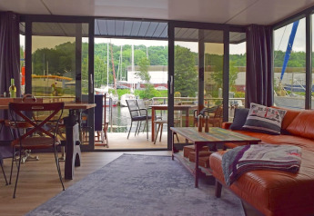 Woonkamer op woonboot ‘Lina’ in Duitsland met grote glazen deuren, uitzicht op de haven en gezellige inrichting.