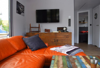 Living area on houseboat 'Lina' in Germany with orange sofa, colorful table, and rustic wooden dresser.