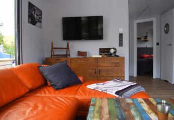 Sala de estar en la casa flotante 'Lina' en Alemania con sofá naranja, mesa colorida y cómoda rústica.