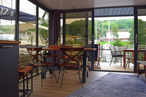 Living area on houseboat «Lina» in Germany with dining table, chairs, and marina view through glass doors.