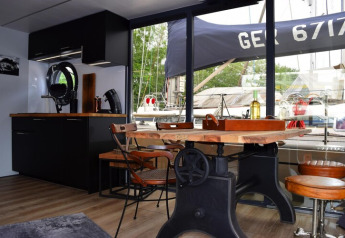 Interior de la casa flotante Lina en Alemania con mesa rústica, cocina moderna y vista al puerto deportivo.