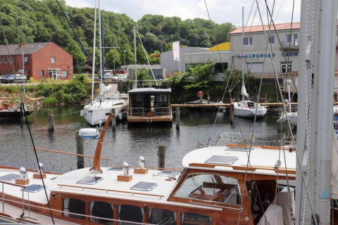 The houseboat 'Lina' is docked in a German harbor, surrounded by sailboats and lush green trees.
