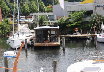 Casa flotante «Lina» atracada en un muelle de Alemania, rodeada de veleros y árboles verdes en verano.