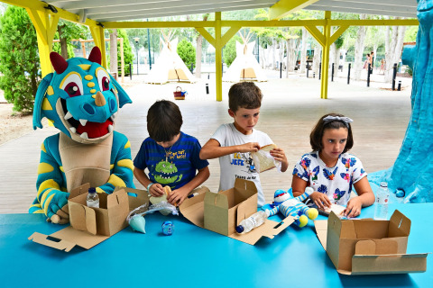 Des enfants et une mascotte dragon bleu font des activités avec des boîtes au Tamarit Park Resort en Espagne.