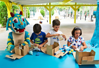 Des enfants et une mascotte dragon bleu font des activités avec des boîtes au Tamarit Park Resort en Espagne.