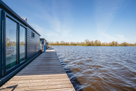Husbåd «Havenlodge Plus» ved Marina Uitgeest i Holland med udsigt over vandet og træer i det fjerne.