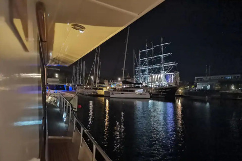 Night view from the houseboat Yacht Aquamarina, looking at illuminated boats and a tall ship in marina.