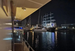 Night view from the houseboat Yacht Aquamarina, looking at illuminated boats and a tall ship in marina.