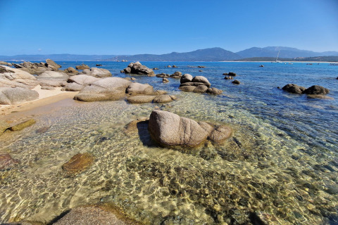 Agua cristalina y rocas en la playa del chalet Résidence U Livanti Belvédère Campomoro, Francia, con montañas.