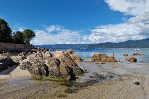 Playa rocosa en Résidence U Livanti Belvédère Campomoro, Francia, con montañas y agua clara en el fondo.