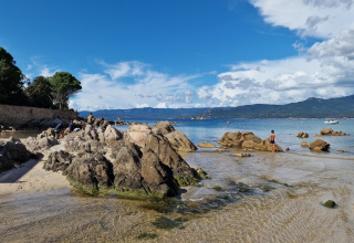 Playa rocosa en Résidence U Livanti Belvédère Campomoro, Francia, con montañas y agua clara en el fondo.