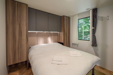 Slaapkamer in Cottage «6p» mobilhome in Frankrijk, tweepersoonsbed, houten kasten en raam met natuurzicht.