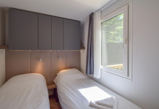 Soveværelse i mobilhome Cottage «6p» i Frankrig med to enkeltsenge, skabe og vindue med gardiner.