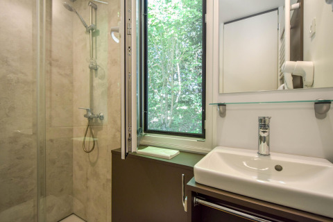 Badkamer met douche, lavabo en open raam met zicht op bomen in het Cottage ‘6p’ mobilhome in Frankrijk.