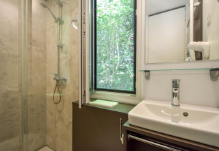 Badkamer met douche, lavabo en open raam met zicht op bomen in het Cottage ‘6p’ mobilhome in Frankrijk.