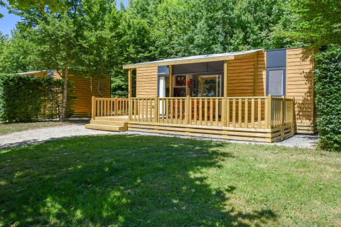 Udendørs billede af en mobilhome Cottage 6p. med træterrasse omgivet af grønne træer og græs.