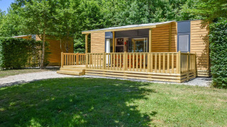 Vista exterior de una casa móvil llamada Cottage 6p. con terraza de madera y rodeada de vegetación.