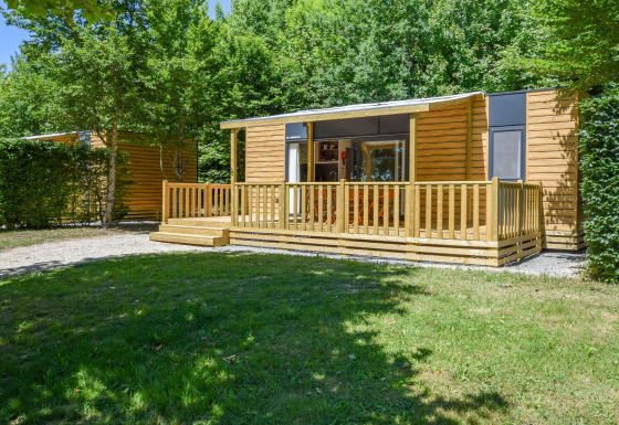 Vista exterior de una casa móvil llamada Cottage 6p. con terraza de madera y rodeada de vegetación.
