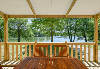 Blick von einer Holzterrasse mit Tisch und Stühlen auf einen See und Bäume am Cottage 6p Mobilheim in Frankreich.