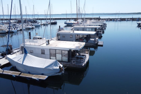 Houseboats docked in a marina alongside sailboats on calm, clear water under a blue sky.