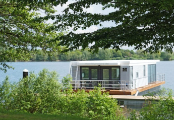Modern houseboat docked on a scenic lake, surrounded by lush greenery and trees on a bright sunny day.