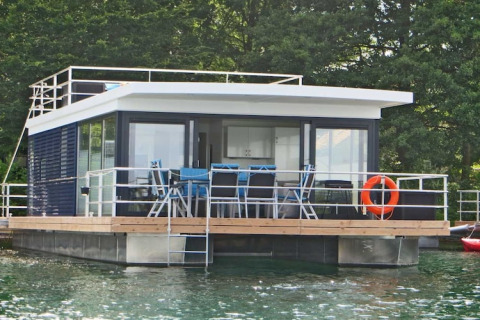 Moderne husbåd Floating 44 med terrasse, liggestole og redningskrans, fortøjet på klart vand, omgivet af grøn skov.
