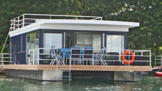 Floating 44 houseboat with deck chairs and a lifebuoy, moored on clear water, surrounded by green trees.