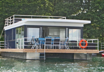 Péniche Floating 44 avec terrasse, chaises et bouée de sauvetage, amarrée sur une eau claire près des arbres.