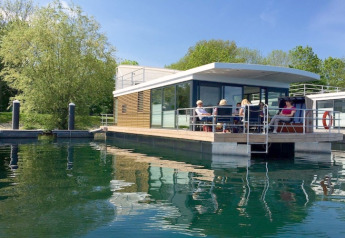Moderne woonboot aangemeerd op rustig water, met mensen die op het dek van een zonnige dag genieten.