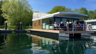 Péniche moderne amarrée sur une eau calme, avec des personnes assises sur le pont profitant du soleil.