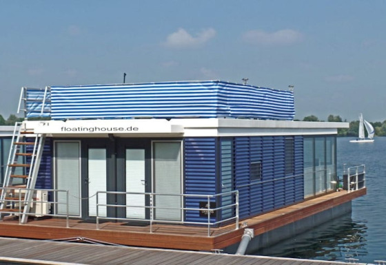 Modernes Hausboot mit blauen und weißen Streifen, Terrasse und Dachleiter auf einem ruhigen See.