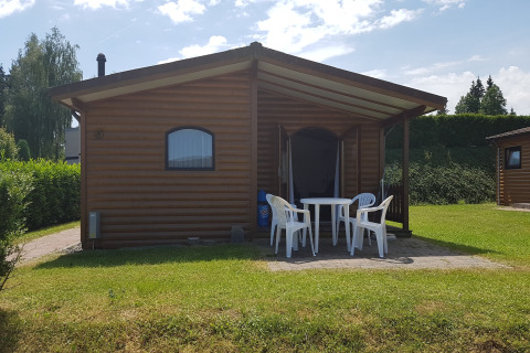 Chalet Waldferienpark Gerolstein 2 met houten buitenkant, witte tuinstoelen en een groen gazon.