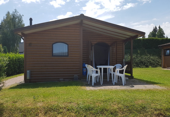 Chalet Waldferienpark Gerolstein 2 met houten buitenkant, witte tuinstoelen en een groen gazon.