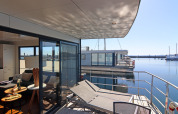 Vista da una moderna casa galleggiante con terrazza e lettini affacciati sull'acqua in una marina soleggiata.