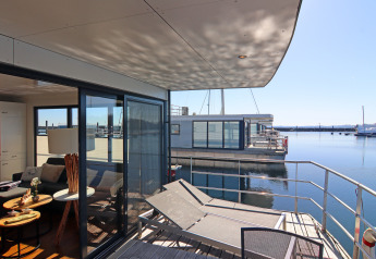 Vista desde una casa flotante moderna con terraza y tumbonas hacia aguas tranquilas en una marina soleada.