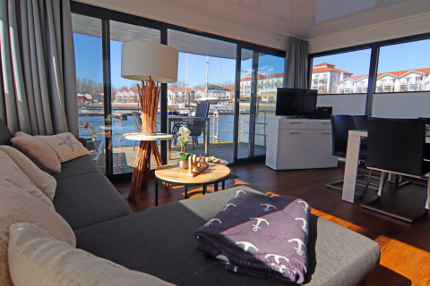 Living area on a houseboat with sofa, tables, TV, and large windows overlooking the waterfront and buildings.