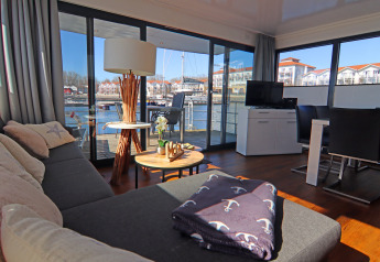 Zona giorno su una houseboat con divano, tavolini, TV e ampie vetrate vista acqua e palazzi circostanti.