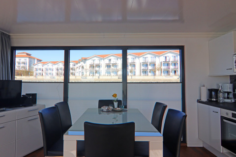 Dining area on a houseboat, featuring large windows with a view of white buildings with red roofs.
