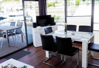 Sala da pranzo moderna su una casa galleggiante con tavolo in vetro, sedie nere, TV e vista sul porto dalle finestre.